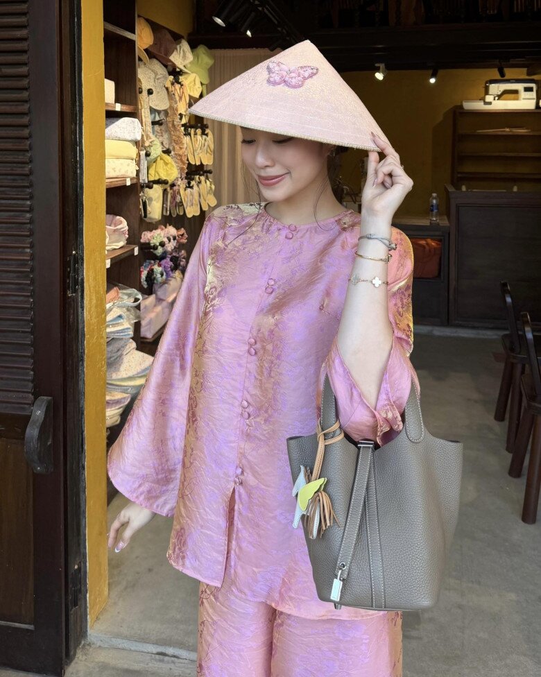 Áo bà ba lụa gấm màu hồng ánh tím, bề mặt vải bắt sáng nhẹ, mang lại vẻ sang trọng kín đáo. Dáng áo suông, tay rộng giúp người mặc thoải mái di chuyển. Phối cùng nón lá cách điệu và túi tote da trung tính, tổng thể vừa giữ nét truyền thống vừa mang hơi thở lifestyle hiện đại.