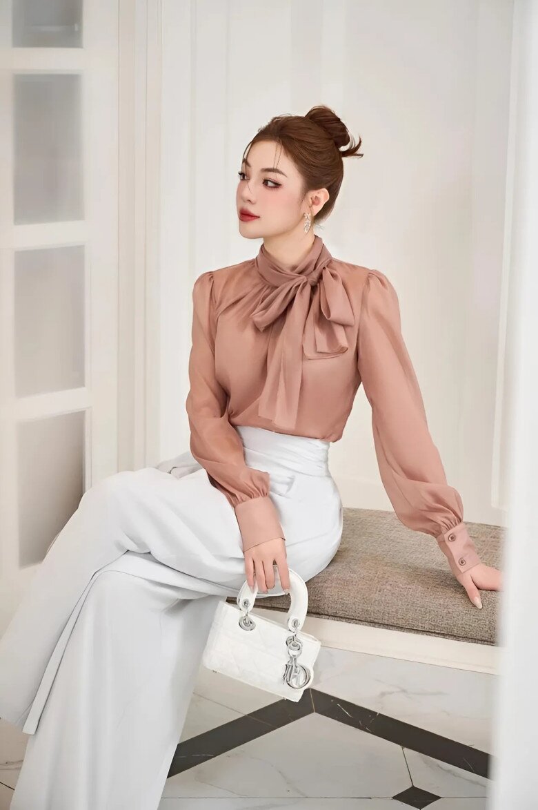 Chất liệu lụa cao cấp ánh nhẹ giúp chiếc áo nâu hồng trở nên sang trọng tuyệt đối. Thiết kế cổ cao đi kèm nơ to bản tạo điểm nhấn nổi bật, tôn lên sự mềm mại mà không kém phần sắc sảo.