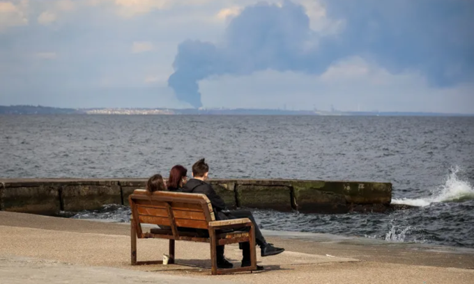 Người dân Ukraine ngồi bên bãi biển nhìn khói bốc lên trên cảng Pivdennyi, sau cuộc tấn công bằng tên lửa của Nga ở Odessa. Ảnh: Reuters