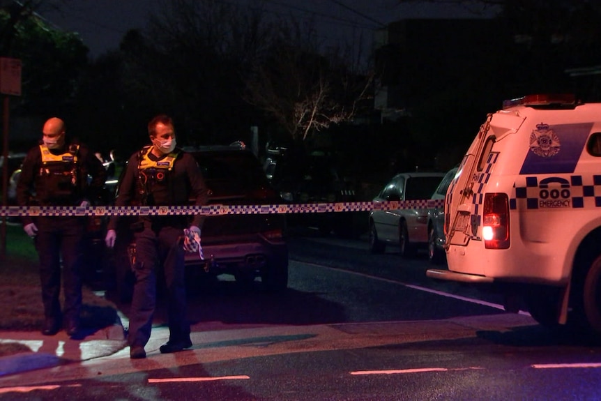 Two police officers behind crime scene tape on a dark street lit by police lights.