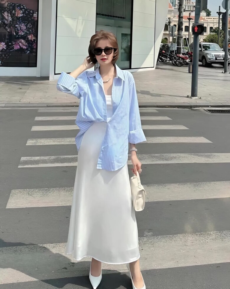 Áo sơ mi sọc xanh phối cùng váy lụa trắng tạo nên tổng thể mềm mại và bay bổng. Cách layering nhẹ nhàng và gam màu pastel khiến set đồ vừa tinh tế vừa hợp với tiết trời hè.
