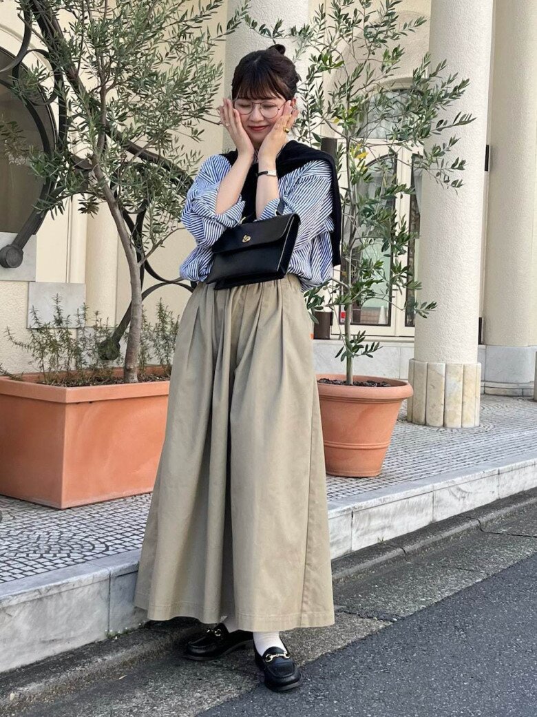 Chiếc áo sơ mi kẻ sọc xanh trắng được sơ vin gọn gàng trong chân váy dài dáng suông màu beige, tạo nên tổng thể nhẹ nhàng, tinh tế. Chiếc áo len khoác hờ trên vai cùng túi xách da đen nhỏ gọn mang lại nét cổ điển kiểu preppy.
