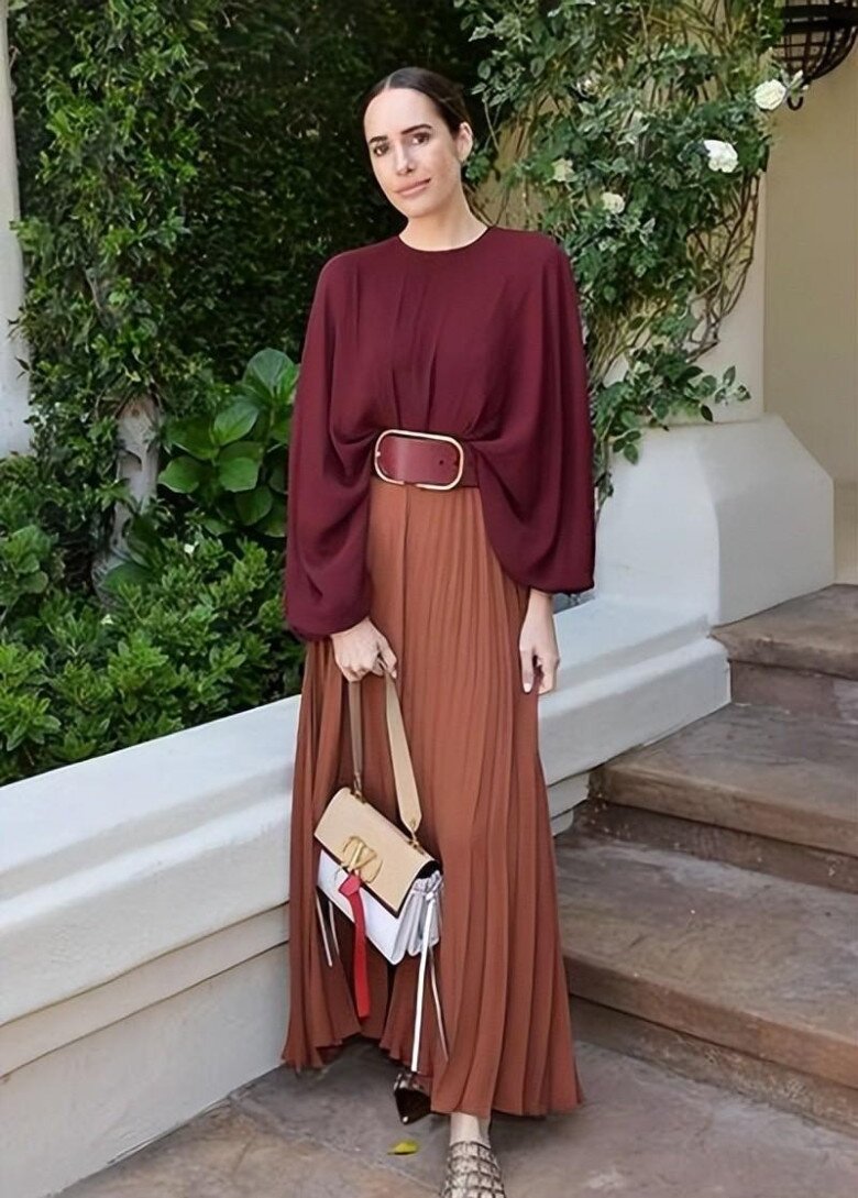 Áo blouse đỏ rượu vang với phần tay phồng mềm mại phối cùng chân váy xếp ly nâu nhạt, tạo nên bố cục chuyển sắc uyển chuyển và đầy chiều sâu. Chiếc thắt lưng bản to cùng túi xách tông nâu be hoàn thiện outfit theo hướng thanh lịch, nữ tính và chuẩn mực “sang trọng không gắng gượng”.