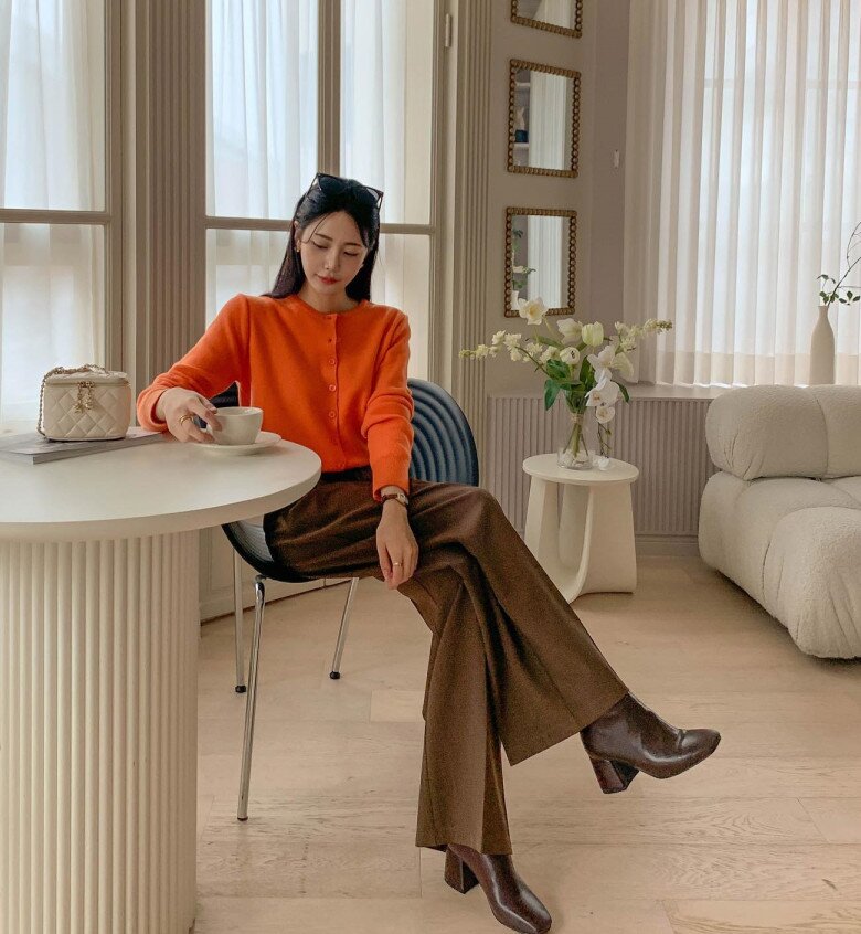 Chiếc áo cardigan cam rực tạo điểm sáng rực rỡ, được tiết chế bởi chiếc quần suông nâu đậm mang hơi hướng retro. Sự phối hợp giữa hai gam màu cùng tông ấm giúp tổng thể vừa nổi bật vừa trầm lắng.