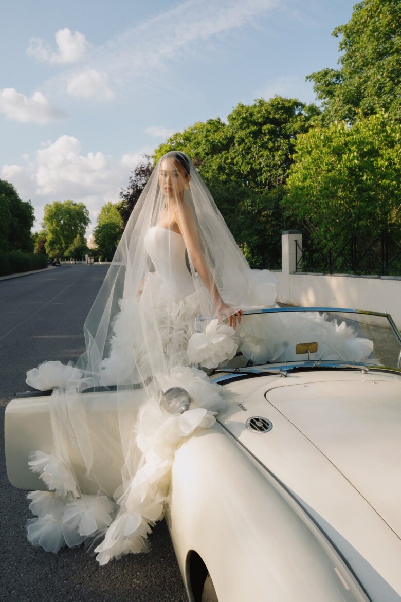 Chiếc váy cưới đầu tiên được cô chọn có phom dáng cúp ngực trơn, ôm sát thân, tạo đường cong mềm mại và tinh giản – xu hướng được nhiều fashion house lớn theo đuổi trong mùa cưới 2025. Phần đuôi váy bồng xòe với những cánh hoa tulle khổng lồ bung nở quanh chân váy, khiến tổng thể vừa cổ điển vừa lãng mạn. Tấm voan dài phủ gần hết chiếc xe cổ càng làm nổi bật vẻ sang trọng mang hơi thở châu Âu của cô dâu.