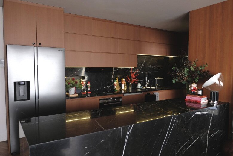 Khu bếp nổi bật trên nền chất liệu gỗ ấm và đá đen sang trọng. Ánh đèn hắt vàng dưới tủ bếp phản chiếu lên nền đá đen giúp các chi tiết đỏ – xanh trở nên nổi bật, đem lại không khí Giáng sinh ấm cúng nhưng hiện đại.