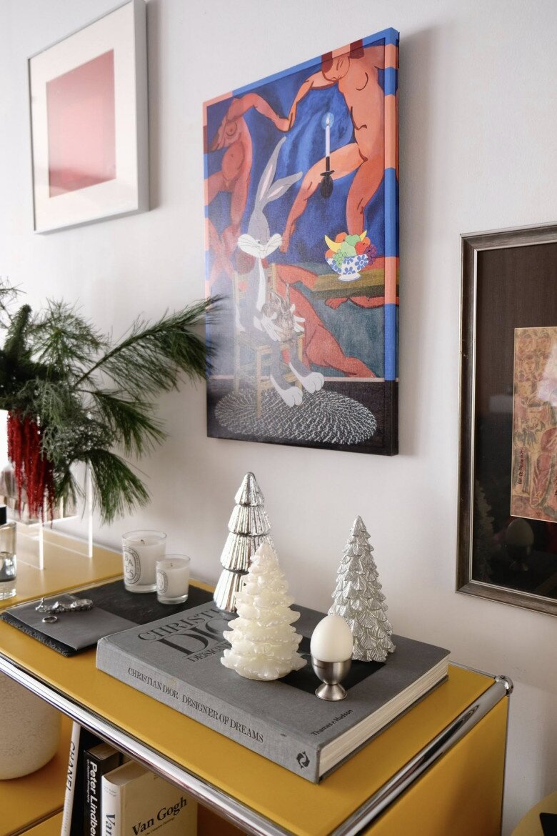 Trên kệ, các vật trang trí nhỏ như nến thơm, bình cành thông tươi, tượng mini hay mô hình cây thông được sắp xếp gọn gàng, còn mảng tường phía sau là những bức tranh màu sắc mạnh từ đỏ, xanh đến trừu tượng khiến góc phòng vừa sinh động vừa nghệ thuật.