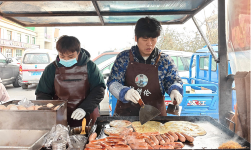 Bai Xianying (bên phải) và vợ làm bánh kếp mặn bên ngoài trung tâm thương mại dệt may ở thành phố Bảo Định, tỉnh Hà Bắc, tháng 2/2026. Ảnh: Cheng Ting/The Paper
